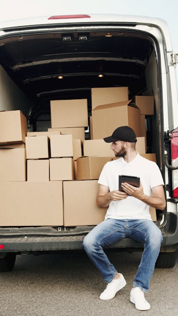 A man with a dark cap and a white polo shirt is standing inside a cargo van filled with neatly stacked cardboard moving boxes of various sizes, some with packing tape, arranged both on the floor and t