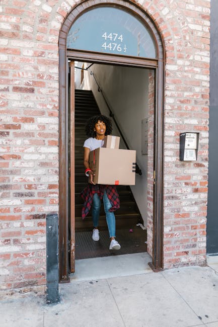 Two movers from Man With a Van Charlton are inside a bright, spacious room with light timber flooring and large arched windows allowing natural daylight to fill the space. One mover, with curly hair and a beard, is standing upright and holding a medium-sized cardboard box at waist height. The second mover, wearing a headband and dressed in dark clothing, is engaged in conversation and holding another cardboard box. Additional boxes are stacked on the floor nearby, some wrapped with red packing tape and others without. The furniture includes a green velvet armchair with wooden legs positioned against the wall on the right. Visible materials in the scene include the cardboard boxes, plastic-wrapped packing tapes, and the clothing of the movers, indicating a moving or home relocation process. This image reflects the packing and loading stages of furniture transport during house removals, with natural lighting highlighting the domestic environment, as part of the services provided by Man With a Van Charlton.