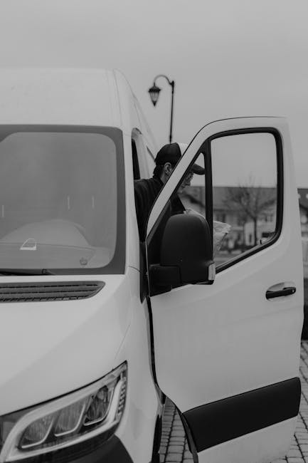 A man wearing a blue beanie and a red and navy jacket stands with arms crossed in front of an open van parked outside a modern building. The van's cargo area is filled with neatly stacked cardboard bo