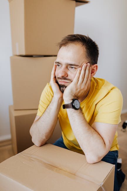 A young man with dark hair and tattoos on his arms is sitting on the wooden floor inside a room, surrounded by cardboard moving boxes of various sizes. He is wearing a black T-shirt and dark trousers 