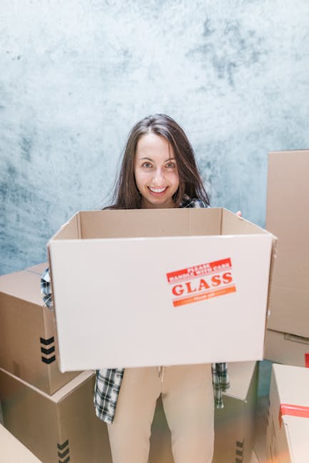 A young woman with curly black hair and a bright smile is sitting on the wooden floor inside an empty room, surrounded by stacked cardboard boxes of various sizes, some with red tape and shipping labe