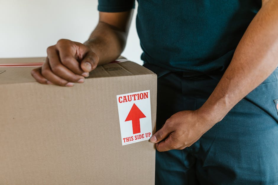 A person wearing a dark green shirt and blue work trousers is holding a large cardboard box with a red and white caution label that reads 'Caution This Side Up' alongside the box, which is inside a property. The individual is gripping the box securely with both hands, preparing for a home relocation. The scene appears to be part of a furniture transport or packing process, with the box positioned on a flat surface, possibly near a doorway or preparing to be loaded into a vehicle. The image emphasizes careful handling and the importance of proper packaging materials during professional removals, illustrating the loading process managed by Man With a Van Charlton as part of their removals service. The background is neutral, and lighting is even, focusing on the actions of loading and careful transport preparation involved in home moving tasks.