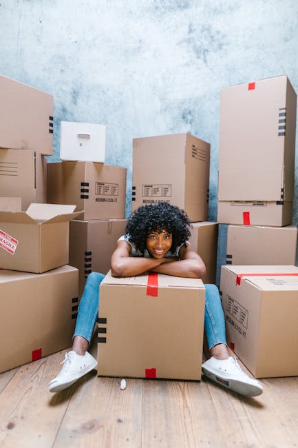 A young woman with curly black hair and a bright smile is sitting on the floor inside a room surrounded by numerous cardboard moving boxes of various sizes, some with printed labels and red tape. She is resting her chin on her arms that are folded over a large box, and her legs are spread out with feet clad in white sneakers. The room has a light blue textured wall and a wooden floor, with natural light illuminating the scene. The boxes are stacked along the walls, indicating an active packing process typical of house relocation and furniture transport preparations. This image, associated with Man With a Van Charlton, illustrates the packing stage of a home move, emphasizing organized handling of packing materials and logistics before actual loading and furniture transport.