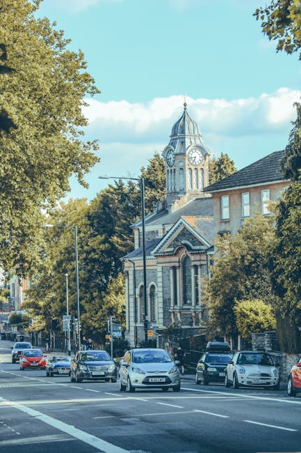 A daytime street scene in Charlton featuring a row of parked cars along the pavement in front of a historic building with a clock tower, which has a domed spire and arched windows, surrounded by tall